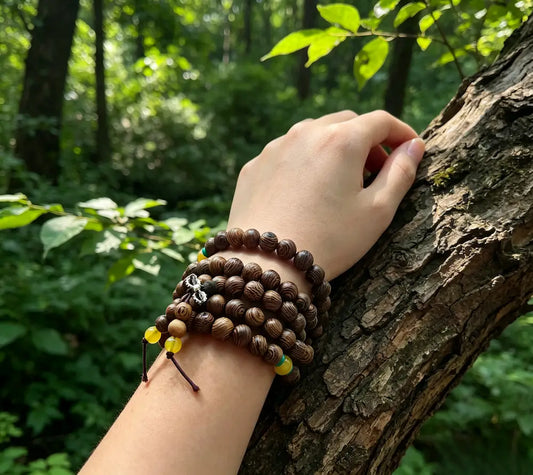 Woman's wrist wearing a 108-bead wenge wood mala bracelet with yellow and turquoise accents, resting on a tree trunk in a green forest with soft natural light.
