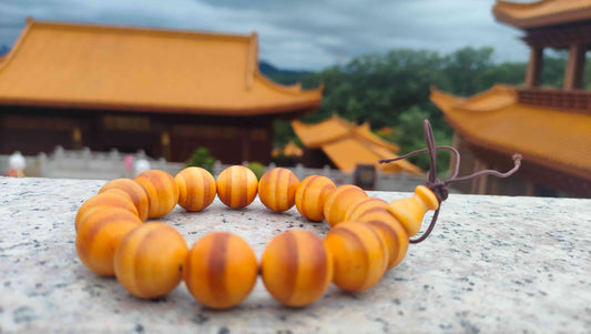 A wooden bead bracelet with tiger-striped grain resting on a temple stone railing, with golden temple roofs and green hills in the background — overcast soft light
