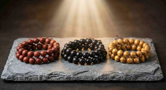 Three natural wood bead bracelets in red, black, and gold colors, neatly arranged on a raw black slate pedestal under a warm top light beam against a dark minimalist background.