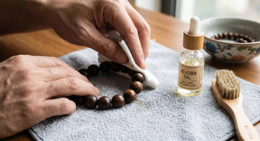 A closeup of hands gently wiping a wenge wood bracelet with a cloth on a gray microfiber towel, next to a glass dropper bottle of jojoba oil and a wooden care brush.
