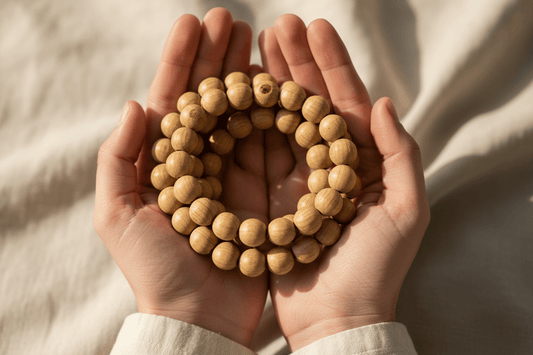  A pair of hands gently holding a light wood mala bracelet against soft neutral fabric.