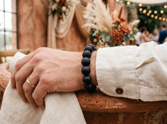  A hand wearing a black wooden bead bracelet and a gold wedding ring, set against soft wedding decor.