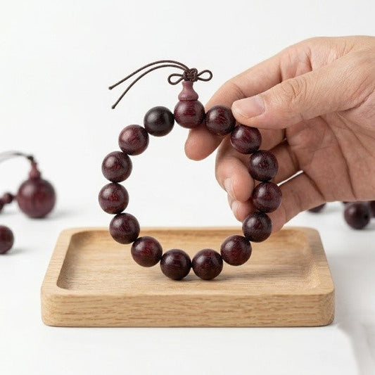  A hand holding a dark red wood bead bracelet above a wooden tray with another bracelet.
