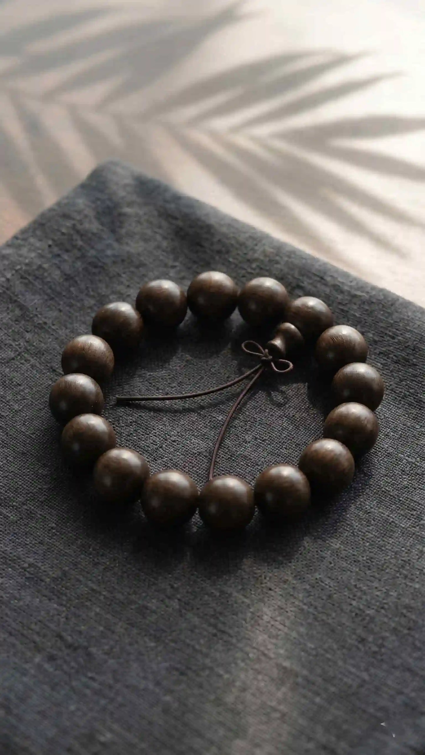 Closeup of an ebony sunken wood bead bracelet with a retro knot, placed on dark gray textured linen fabric with soft plant leaf shadows.
