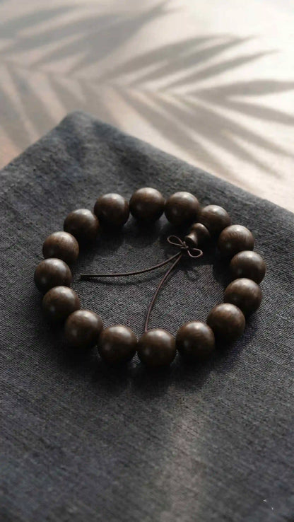 Closeup of an ebony sunken wood bead bracelet with a retro knot, placed on dark gray textured linen fabric with soft plant leaf shadows.