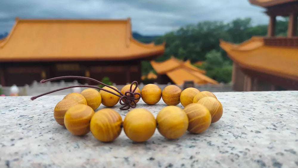 A golden sandalwood bead bracelet with a brown knot on a marble railing, with traditional oriental temple architecture and golden roofs in the background.