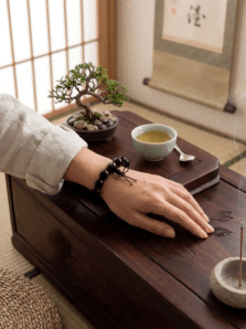 A serene, Zen-inspired setting featuring a hand wearing a Lumala black ebony gourd bracelet resting on a rustic dark wooden tea table. In the background, a small bonsai tree, a traditional tea set, and a Japanese shoji screen enhance the mindful living aesthetic.
