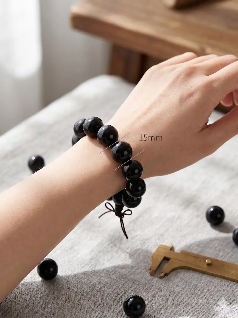 Close-up of a 15mm natural black ebony wood bead meditation bracelet worn on a wrist, with elegant overlay text describing the balanced 15mm statement size and premium craftsmanship.
