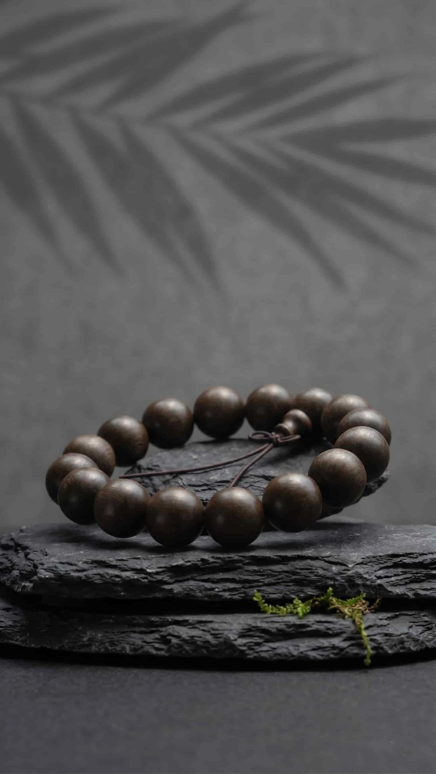 Oriental Zen style ebony wood prayer beads displayed on a raw black stone pedestal with green moss, set against a dark gray wall with tree silhouettes.
