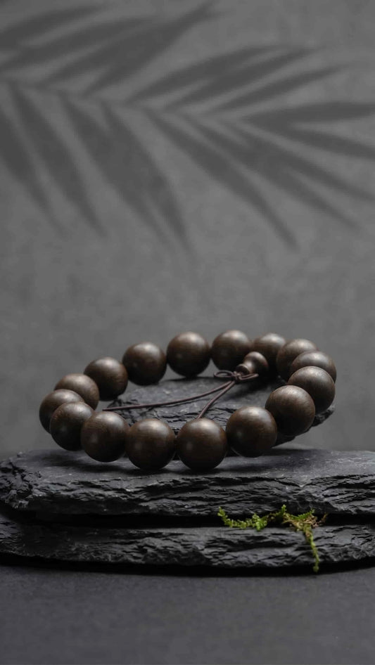 Oriental Zen style ebony wood prayer beads displayed on a raw black stone pedestal with green moss, set against a dark gray wall with tree silhouettes.