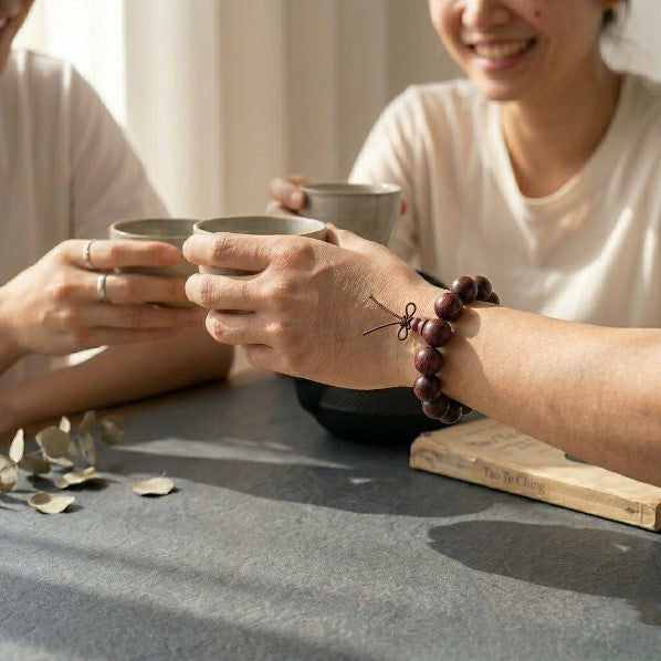 A close-up of a person wearing a 15mm rosewood mala beads bracelet on their wrist, holding a ceramic teacup during a social gathering. A vintage Tao Te Ching book and dried leaves sit on a dark slate table under warm, gentle sunlight.