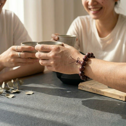 A close-up of a person wearing a 15mm rosewood mala beads bracelet on their wrist, holding a ceramic teacup during a social gathering. A vintage Tao Te Ching book and dried leaves sit on a dark slate table under warm, gentle sunlight.