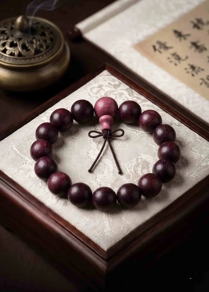 A premium 15mm rosewood mala beads bracelet placed neatly on a luxurious cream-colored silk padded wooden display box. In the background, a bronze incense burner emits soft white smoke next to a traditional Asian calligraphy scroll.