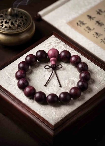A premium 15mm rosewood mala beads bracelet placed neatly on a luxurious cream-colored silk padded wooden display box. In the background, a bronze incense burner emits soft white smoke next to a traditional Asian calligraphy scroll.
