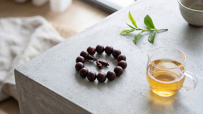 A top-down shot of a rich burgundy wood bead bracelet next to a glass teacup filled with vibrant golden tea, accented by fresh green leaves under bright, clear light.
