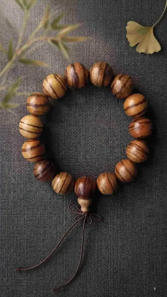 Top-down closeup of a wenge wood bead bracelet with chicken-wing feathered stripes, placed on textured charcoal gray linen under soft 