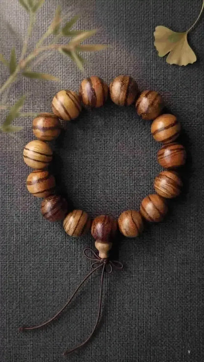 Top-down closeup of a wenge wood bead bracelet with chicken-wing feathered stripes, placed on textured charcoal gray linen under soft 