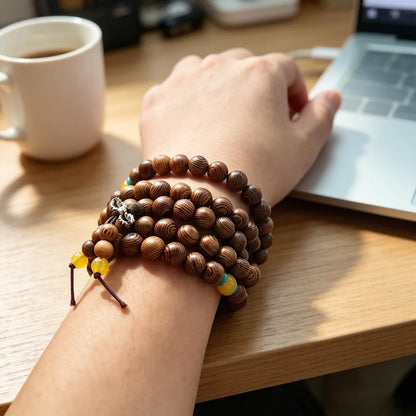 Hand wearing a multi-wrap wenge wood 108-bead mala bracelet with yellow accents, resting near a coffee cup and laptop on a wooden table.

