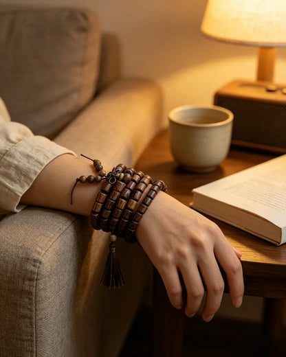 Natural wenge feather grain mala bracelet worn on wrist, warm living room night scene with sofa, reading book and table lamp, quiet mindfulness accessory