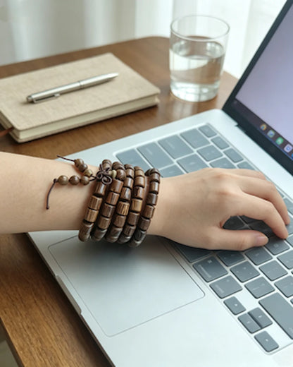 Wenge wood barrel bead mala bracelet wrapped four times on wrist, daily office work scene with silver laptop, notebook and water glass, natural feather grain wooden meditation bracelet for Lumala brand